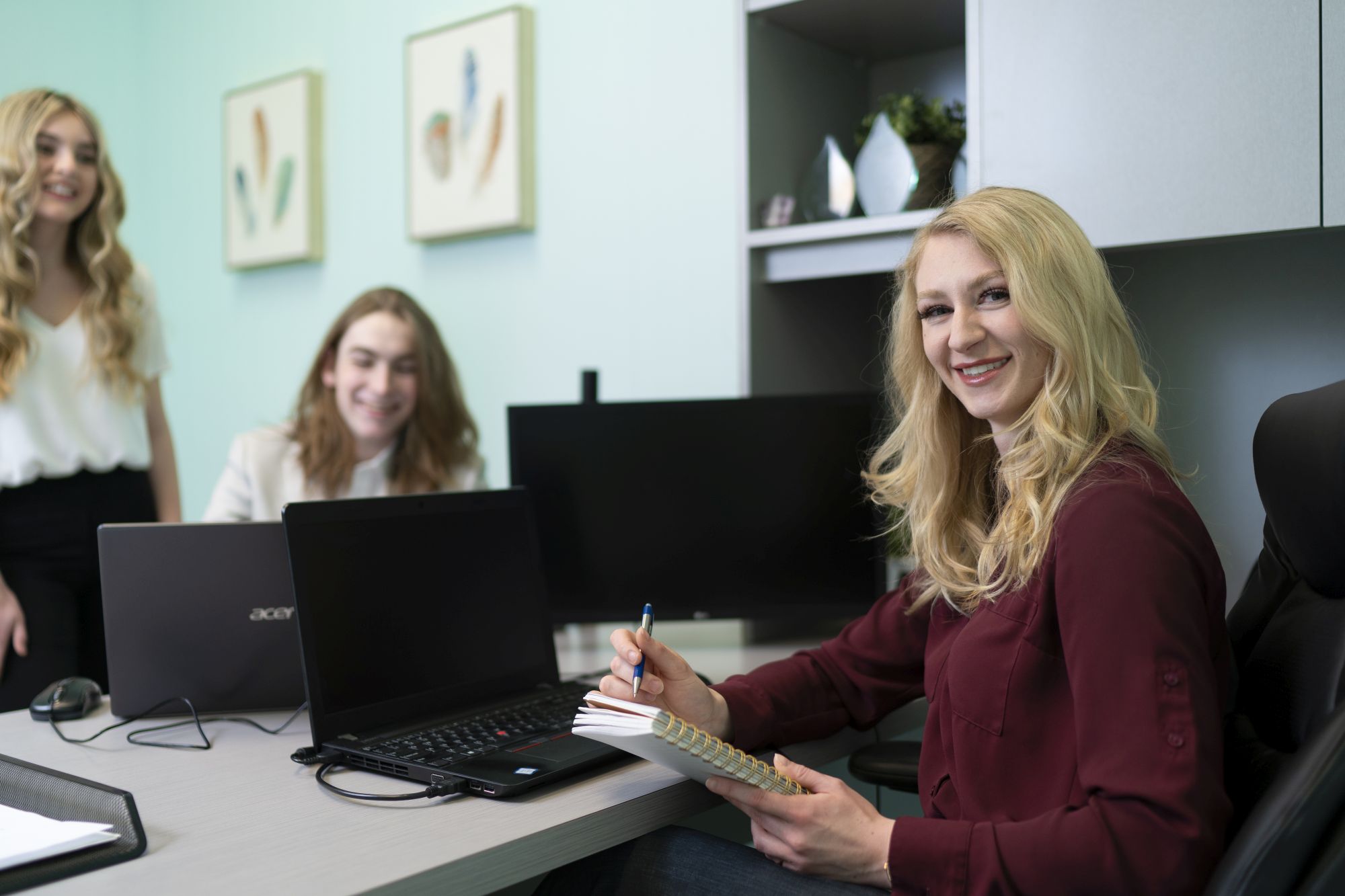 A student advisor smiling and taking notes while meeting with two students at a desk with laptops in a MaKami Education Foundation office in Alberta